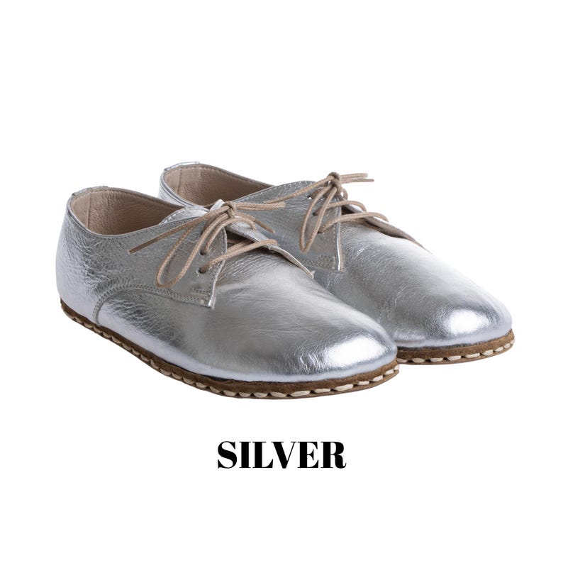 May include: A pair of metallic silver lace-up shoes with a rounded toe, low profile, and light tan laces. The shoes feature visible stitching along the sole edge and a smooth, reflective finish. The word SILVER is printed in black below the shoes.