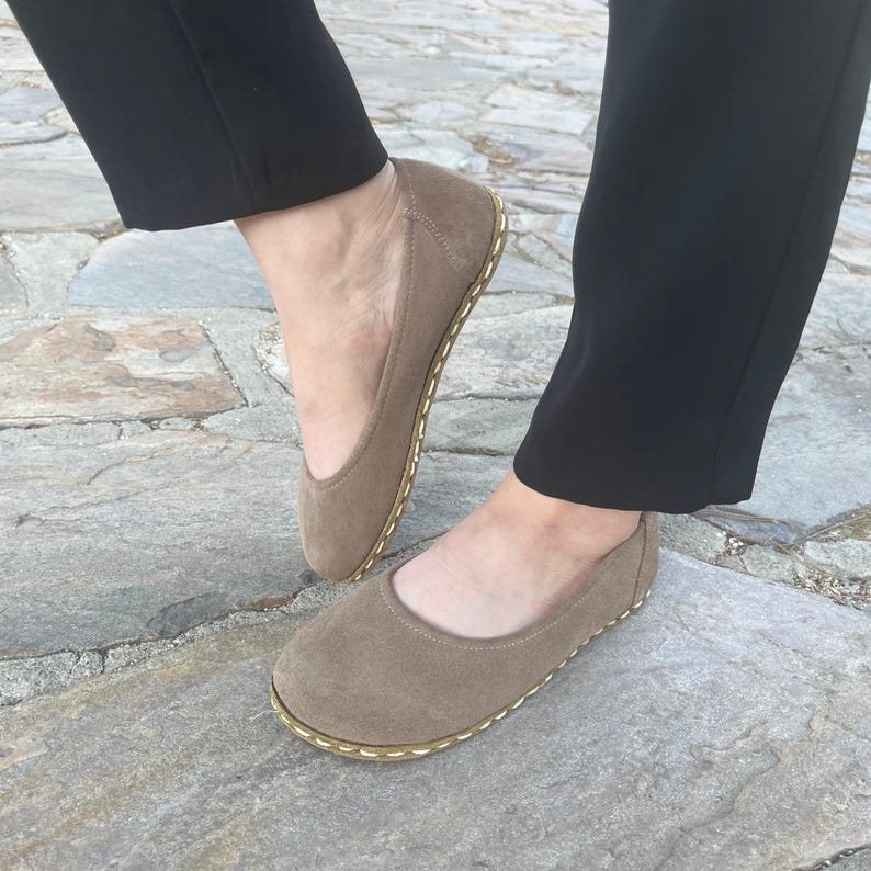 May include: Taupe suede ballet flats with a rounded toe and a stitched sole. The flats are worn with black pants. The shoes have a minimalist design, suitable for casual wear.
