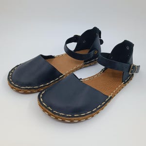 May include: A pair of dark navy blue flat sandals featuring a rounded toe, an ankle strap with a small metal buckle, and a tan textured insole. The outer edge of the sole is finished with a thick, visible light-coloured whipstitch detail.