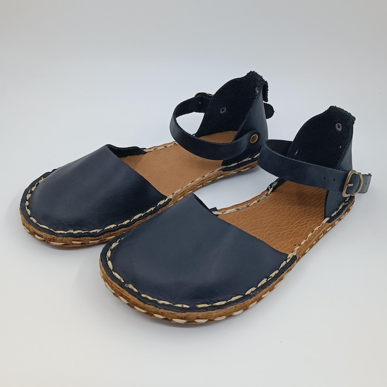 May include: A pair of dark navy blue flat sandals featuring a rounded toe, an ankle strap with a small metal buckle, and a tan textured insole. The outer edge of the sole is finished with a thick, visible light-coloured whipstitch detail.