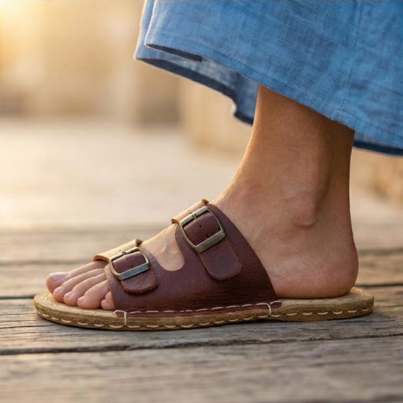 May include: Brown leather sandals with two adjustable straps and metal buckles. The sandals have a cork footbed and are stitched around the edges. The person is wearing a blue garment.