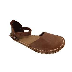 May include: A brown leather sandal featuring a closed toe, an open heel, and an ankle strap with a metal buckle. The sole is attached to the upper with visible white stitching that runs along the entire perimeter of the shoe's base.