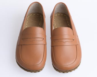 May include: A pair of tan leather loafers featuring a rounded toe, a classic penny slot strap across the vamp, and visible light-coloured stitching along the sole edge. The interior of the shoes shows a tan lining with a stitched insole detail.