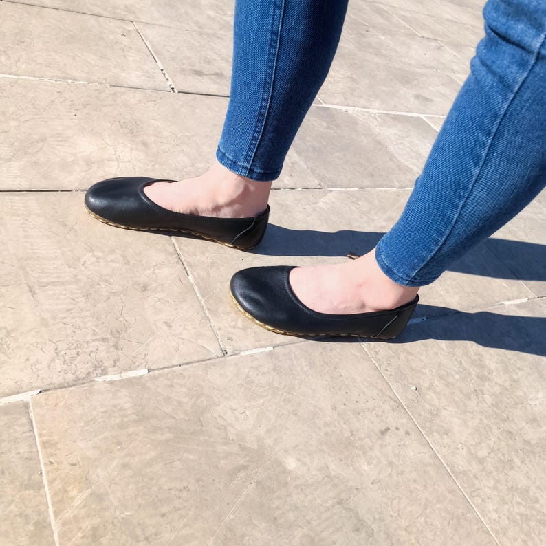 May include: A pair of black leather ballet flats with a gold stitching detail. The flats are worn by a person wearing blue jeans.