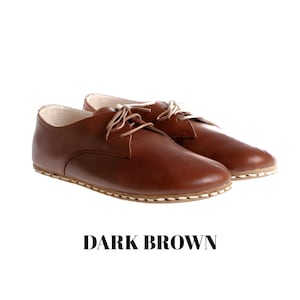 May include: A pair of dark brown leather shoes with a low-cut profile, rounded toes, and thin beige laces. The shoes feature visible light-coloured stitching along the sole edge and a smooth finish. The text DARK BROWN is printed below the shoes.