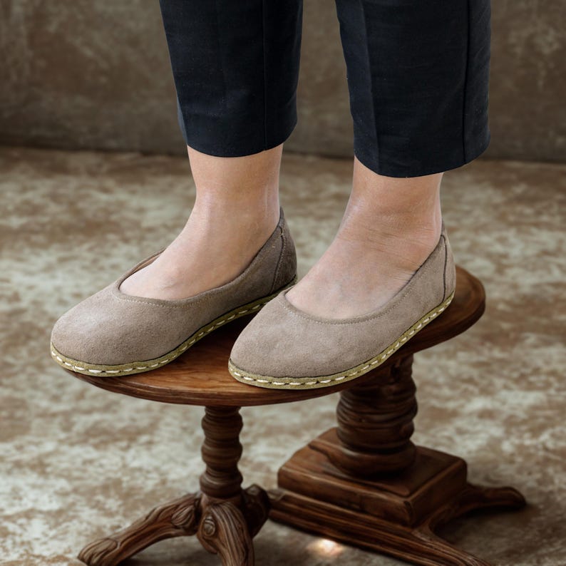 May include: A pair of taupe-colored ballet flats with a rounded toe and a stitched sole. The shoes are made of suede-like material and are worn by a person wearing black pants. The shoes are on a small, ornate wooden table.