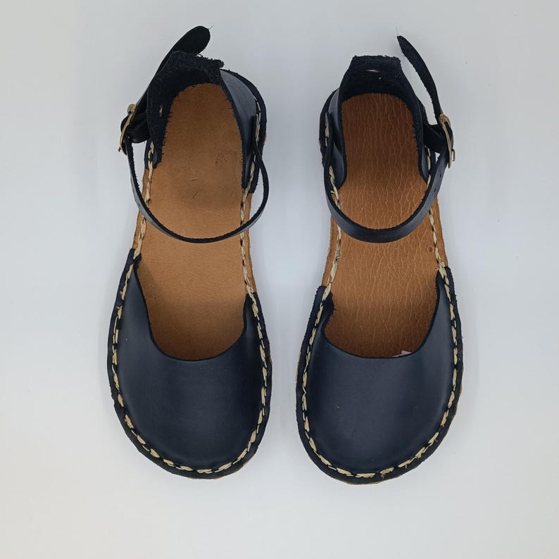 May include: A pair of black leather sandals featuring a rounded toe, an ankle strap with a metal buckle, and a tan-coloured inner sole. The outer edges of the sandals are finished with a visible, thick, light-coloured whipstitch detail along the sole.