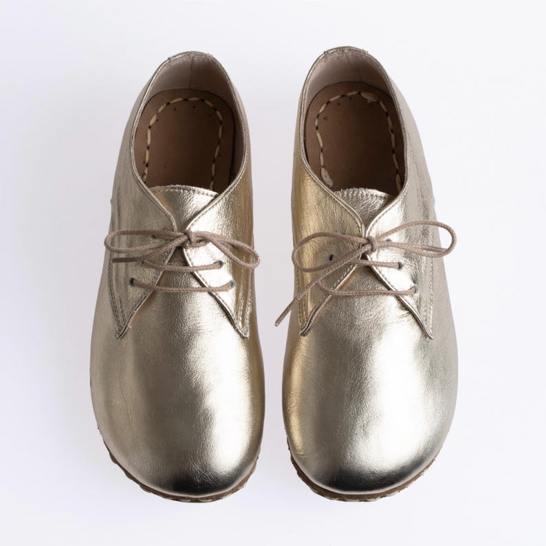 May include: A pair of metallic gold leather shoes featuring a rounded toe, a low-cut ankle profile, and thin beige laces threaded through two eyelets on each side. The interior lining is tan with visible stitching along the inner edge of the sole.
