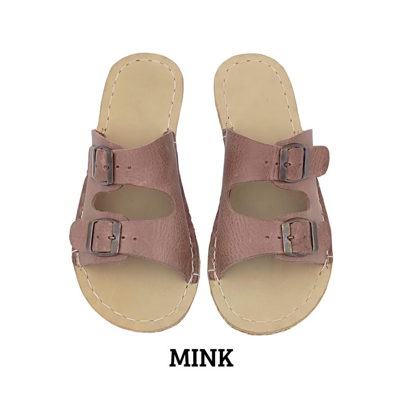 May include: A pair of mink-colored leather sandals with two adjustable buckle straps. The sandals have a light tan sole and are stitched with white thread. The word "MINK" is printed in bold, black letters below the sandals.
