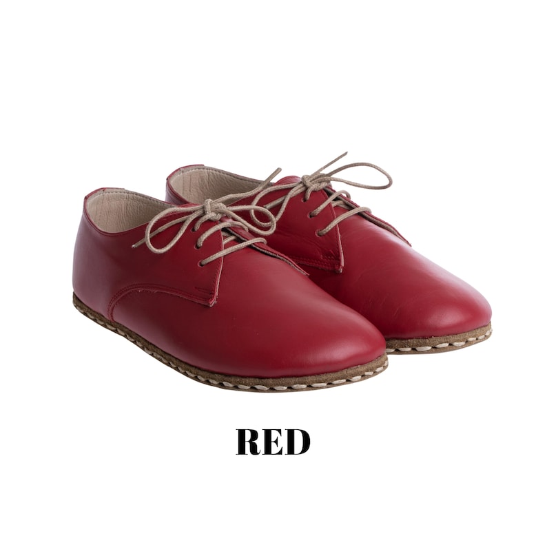 May include: A pair of red leather oxford shoes with beige laces and visible stitching along the sole. The shoes feature a smooth finish and a rounded toe design. The word RED is printed in bold black capital letters below the shoes.