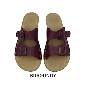 May include: Burgundy leather sandals with two adjustable straps and metal buckles. The sandals have a light brown footbed and white stitching around the edges. The word "BURGUNDY" is printed in bold, black letters below the sandals.