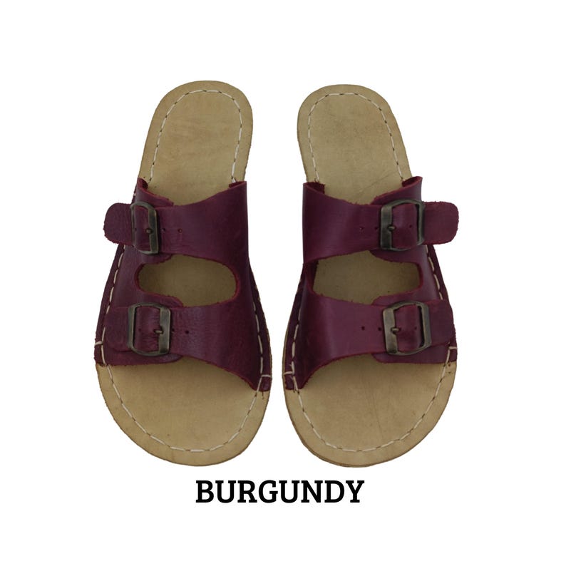May include: Burgundy leather sandals with two adjustable straps and metal buckles. The sandals have a light brown footbed and white stitching around the edges. The word "BURGUNDY" is printed in bold, black letters below the sandals.