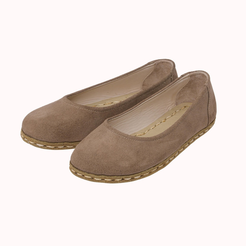 May include: A pair of taupe-colored suede ballet flats. The shoes have a rounded toe and a stitched sole. The interior is a lighter shade, and the overall design is simple and classic. These flats are a versatile footwear option.