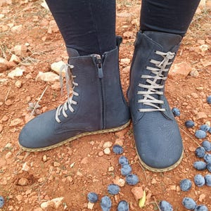 May include: A pair of dark blue leather boots with laces and a zipper. The boots have a thick sole and are worn on a dirt path with blue berries scattered around.
