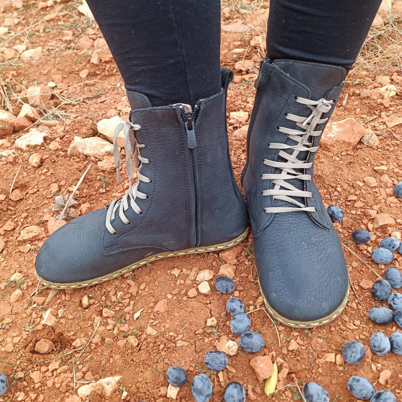 May include: A pair of dark blue leather boots with laces and a zipper. The boots have a thick sole and are worn on a dirt path with blue berries scattered around.