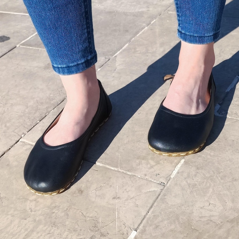 May include: A pair of black leather ballet flats with gold trim around the edges. The flats are worn by a person wearing blue jeans.