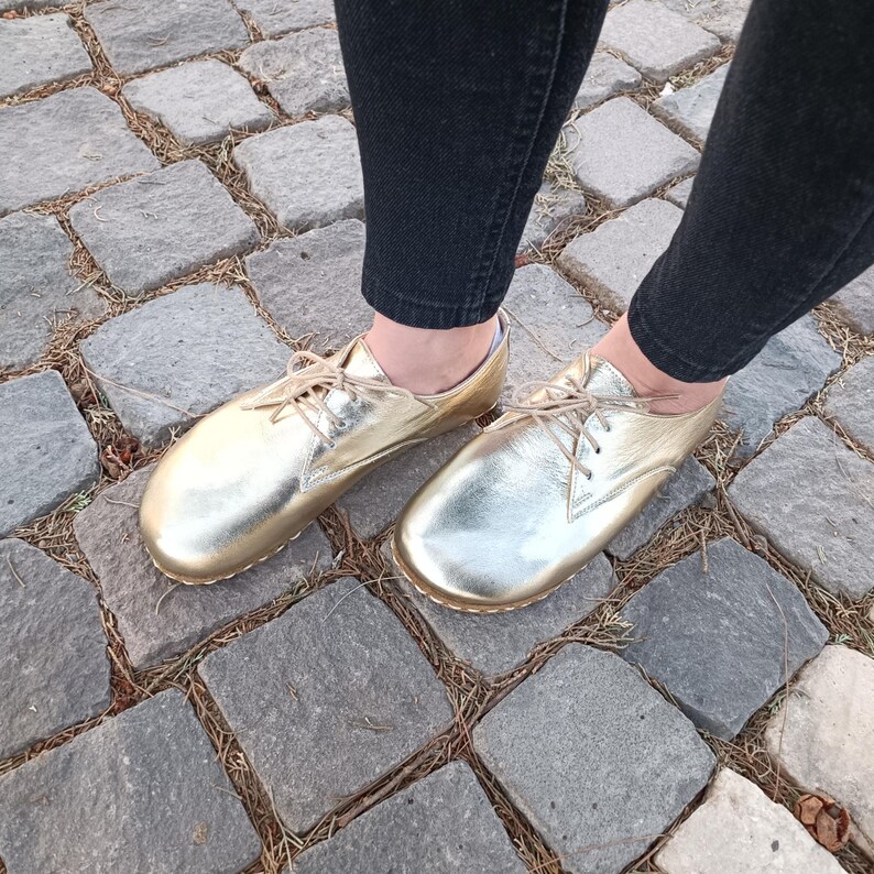 May include: A pair of metallic gold leather oxford shoes with thin matching laces and a rounded toe design. The shoes feature a smooth, reflective finish and are worn with dark denim jeans, showcasing a low-profile silhouette and stitching.