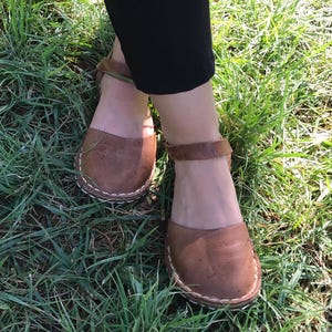 May include: A pair of brown leather sandals featuring a closed-toe design with a rounded front, an ankle strap with a buckle closure, and visible white stitching along the sole edge, worn with black trousers.