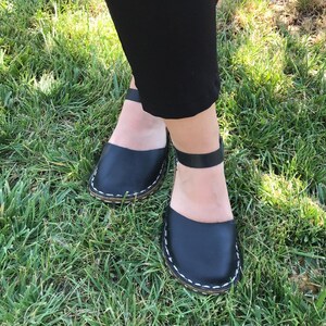 May include: A pair of black leather sandals featuring a wide front strap and a thin ankle strap with a buckle closure. The edges of the sandals are detailed with visible white stitching along the sole, providing a contrast to the black leather.