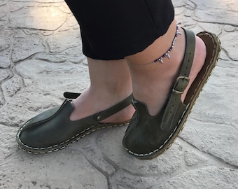 May include: A pair of olive green leather sandals with a strap and buckle closure. The sandals have a white stitching detail around the edges and a rubber sole.