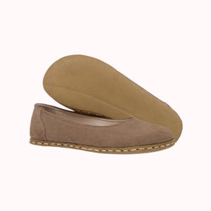 May include: A pair of taupe-colored ballet flats with a suede-like texture. The sole is a light brown color, and the shoe is stitched with a contrasting thread around the edge. One shoe is positioned above the other, showing the sole.