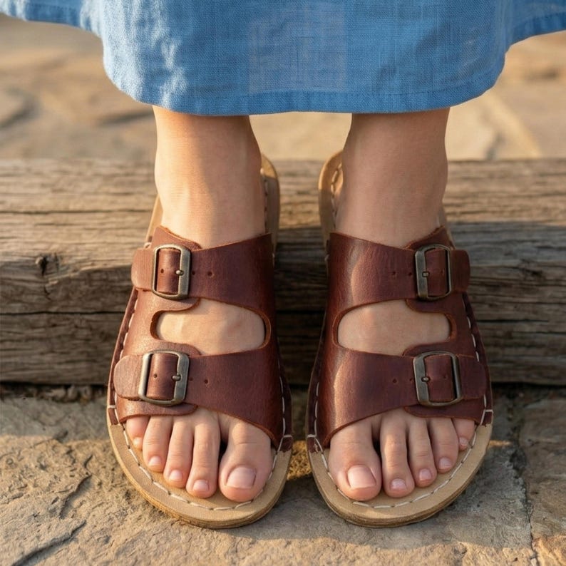 May include: Brown leather sandals with two adjustable straps and metal buckles. The sandals have a light-colored sole and are stitched with white thread. The image shows the sandals on a wooden surface.