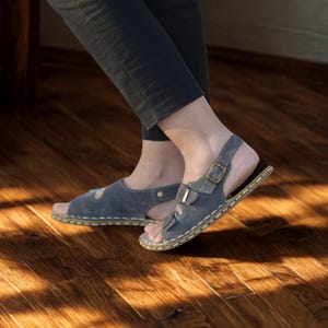 Può includere: Sandali in pelle grigia con punta aperta e cinturini alla caviglia regolabili. I sandali hanno una chiusura con fibbia e un design cucito sui bordi. Le suole sono di colore marrone chiaro.