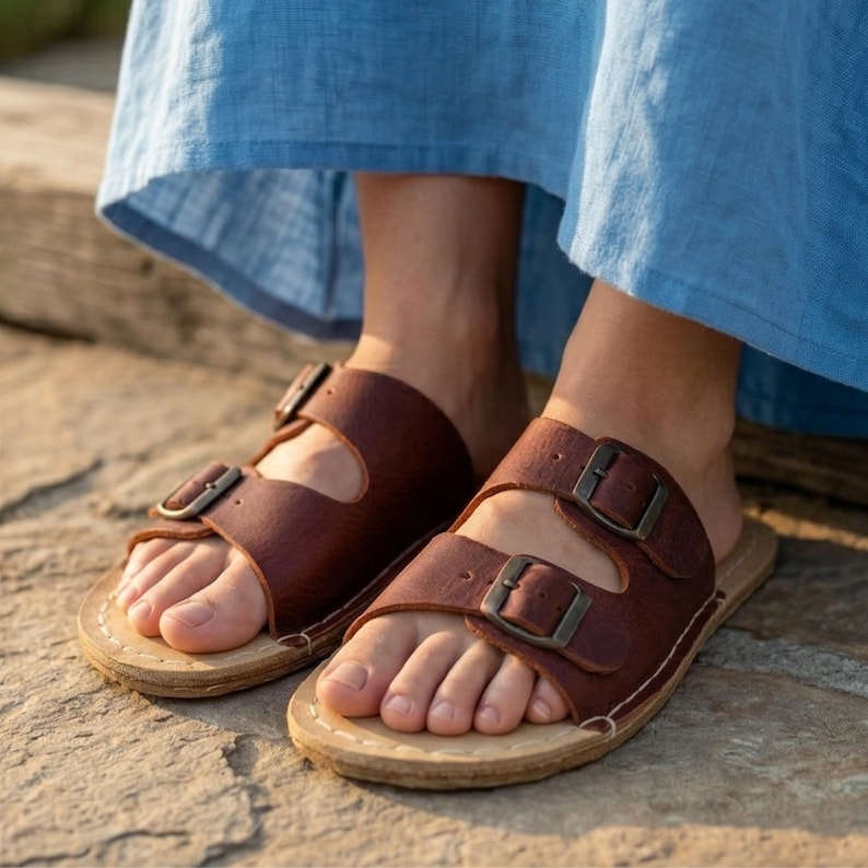 May include: Brown leather sandals with two adjustable straps and metal buckles. The sandals have a light-colored sole and are worn by a person wearing a blue dress. The sandals are designed for comfort and style.