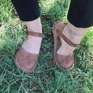 May include: A pair of brown leather sandals featuring a rounded toe, a single ankle strap with a side buckle, and visible white stitching along the sole edge. The sandals have a flat construction with a textured leather finish and a simple design.