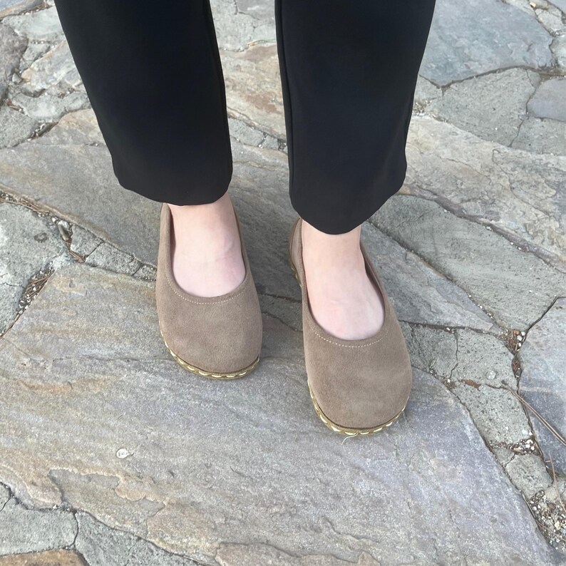 May include: A pair of taupe-colored ballet flats. The shoes have a rounded toe and a thin, gold-colored trim around the sole. The shoes are made of a soft material and are worn with black pants.