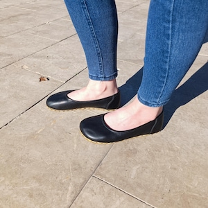 May include: A pair of black leather ballet flats with a gold trim around the sole. The flats are worn by a person wearing blue jeans.