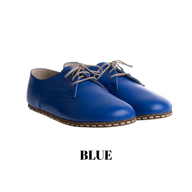 May include: A pair of vibrant blue lace-up shoes featuring a smooth finish, light-coloured laces, and visible stitching along the sole edge. The word BLUE is printed in bold black capital letters below the shoes on a plain white background.