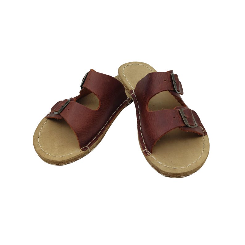 May include: A pair of brown leather sandals with two adjustable straps and metal buckles. The sandals have a light tan footbed and are stitched with white thread. The footwear is designed for casual wear.