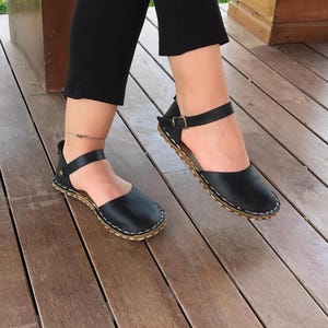 May include: A pair of black leather sandals featuring a rounded toe, an ankle strap with a metallic buckle, and a visible light-coloured stitched edge along the sole. The design includes a heel strap and a smooth finish on the upper leather material.