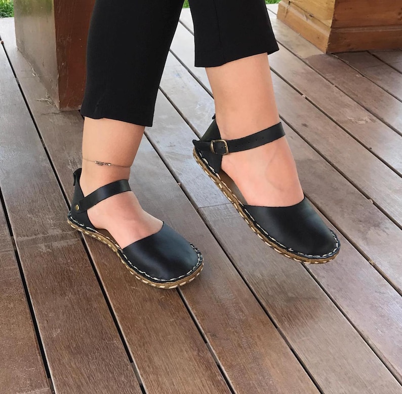 May include: A pair of black leather sandals featuring a rounded toe, an ankle strap with a metallic buckle, and a visible light-coloured stitched edge along the sole. The design includes a heel strap and a smooth finish on the upper leather material.
