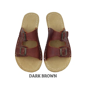 May include: A pair of dark brown leather sandals with two adjustable buckle straps. The sandals have a light tan insole and are stitched with white thread. The text "DARK BROWN" is displayed below the sandals.