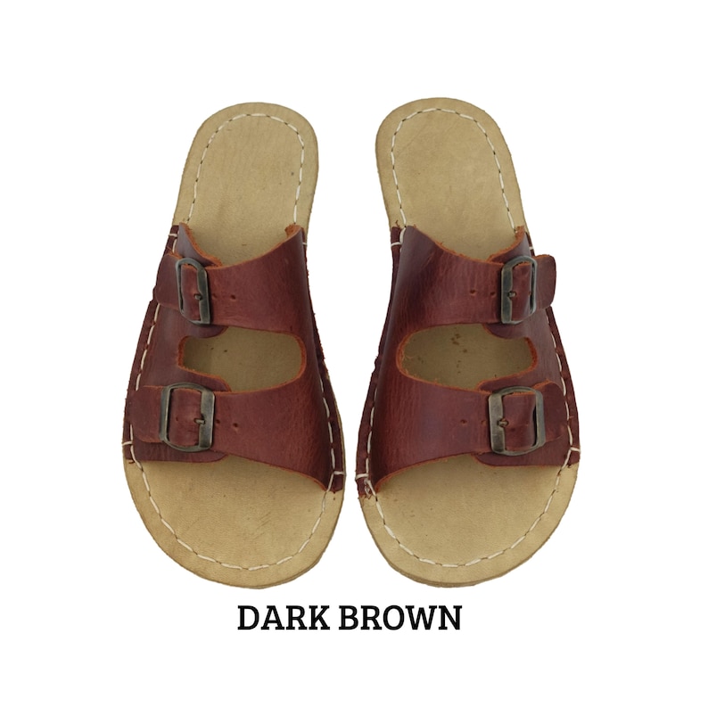 May include: A pair of dark brown leather sandals with two adjustable buckle straps. The sandals have a light tan insole and are stitched with white thread. The text "DARK BROWN" is displayed below the sandals.
