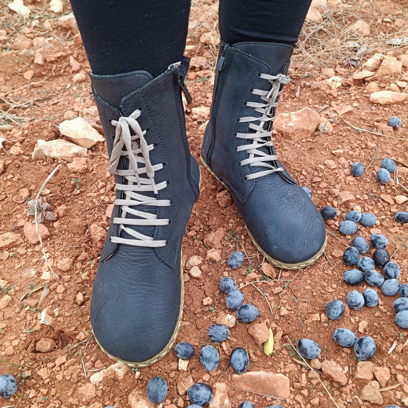 May include: A pair of black leather ankle boots with gray laces. The boots are worn and have a distressed look. The boots are on a dirt path with blue berries scattered around them.