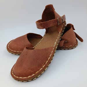 May include: A pair of brown leather sandals featuring a closed-toe design, an ankle strap with a metal buckle, and visible white whipstitching along the edges of the sole. The leather has a textured, matte finish with a natural appearance.