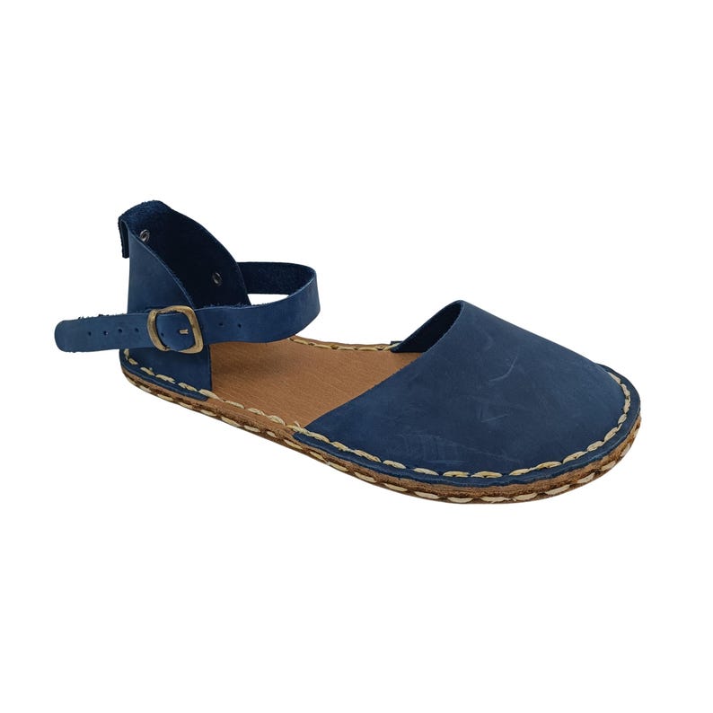 May include: A blue flat sandal featuring a closed toe, an ankle strap with a bronze-toned metal buckle, and a tan-colored footbed. The sole is attached to the upper with visible light-colored stitching that runs along the entire perimeter.