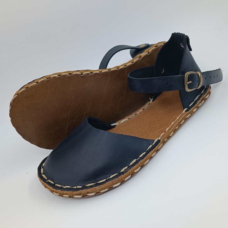 May include: A pair of dark navy blue leather sandals featuring a rounded toe, an ankle strap with a brass-toned metal buckle, and a tan leather insole. The upper is stitched to the sole with visible light-coloured thread along the outer edge.