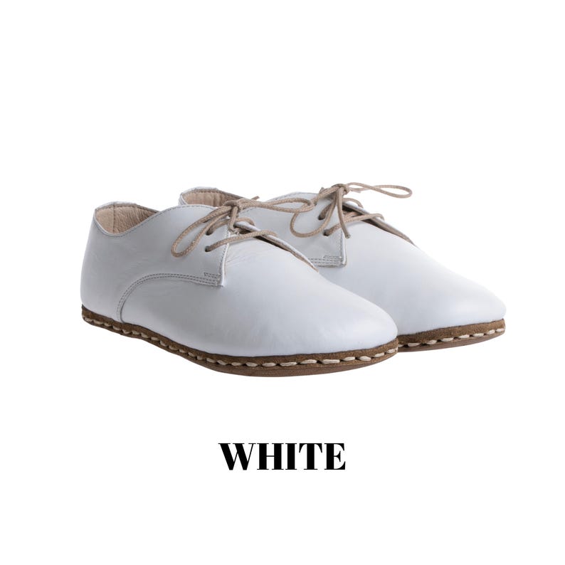 May include: A pair of white leather lace-up shoes featuring a rounded toe, beige laces, and a visible brown stitched welt along the sole. The word WHITE is printed in bold black capital letters below the shoes on a plain white background.