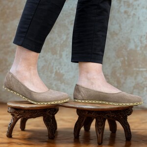 May include: Tan suede ballet flats with a rounded toe and stitched sole. The shoes are being worn, with the wearer's legs in black pants. The shoes are on small, ornate wooden stools.