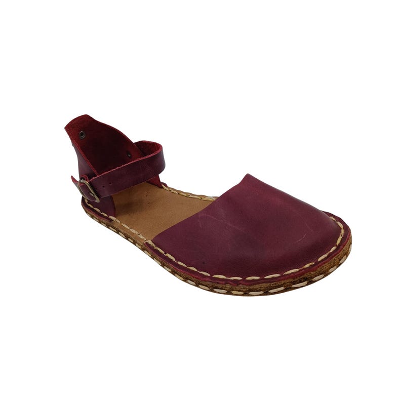 May include: A deep burgundy leather sandal featuring a rounded toe, a wide front strap, and an ankle strap with a small metal buckle. The sole is outlined with thick, visible white stitching that connects the upper leather to the tan inner sole.