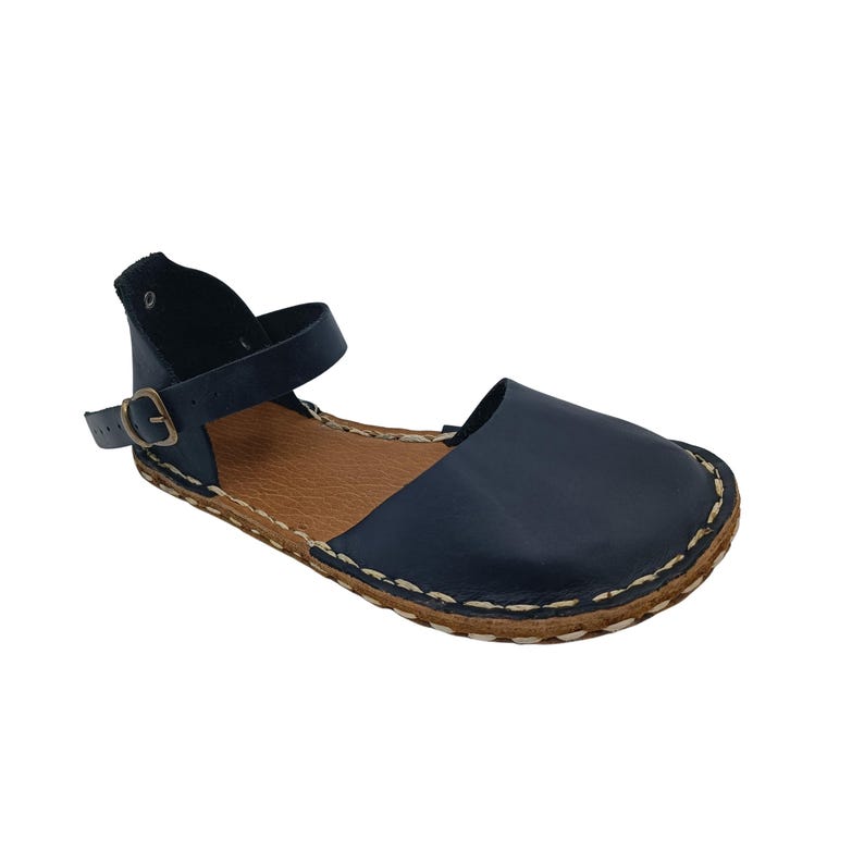 May include: A dark navy blue leather sandal featuring a wide front strap and an adjustable ankle strap with a small bronze-toned metal buckle. The sole is tan with visible light-colored stitching along the edge, connecting the upper to the base.