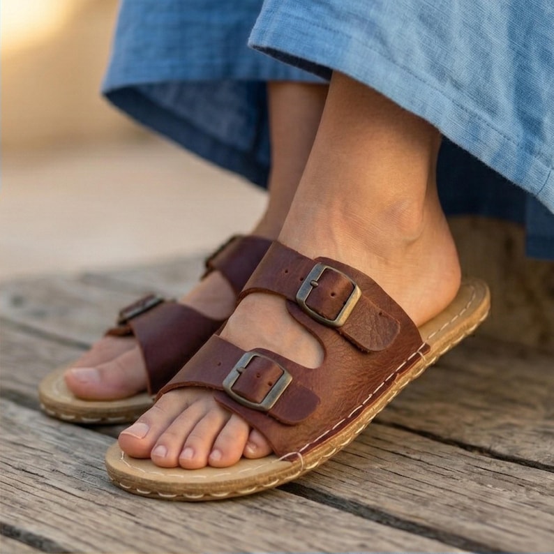 May include: Brown leather sandals with two adjustable straps and metal buckles. The sandals have a light-colored sole and are designed for comfort and style. Perfect for casual wear.