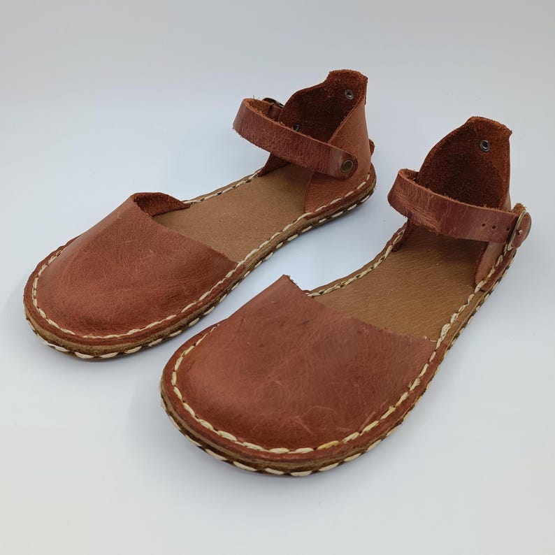 May include: A pair of brown leather sandals featuring a rounded toe, an ankle strap with a metal buckle, and visible light-colored stitching along the perimeter of the sole. The sandals have a flat construction with a textured leather finish.