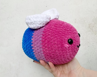 May include: A crocheted plush toy shaped like a bee made with blue, pink and white yarn. The bee has a white crocheted wing on its back and black eyes.