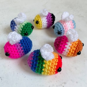 May include: Six crocheted bumblebees in various colours, including rainbow, pink, blue, and green. Each bee has white wings and black eyes.