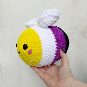 May include: A round, plush crochet bee toy featuring horizontal stripes of yellow, white, purple, and black yarn. The bee has two white textured wings on top, two round black plastic eyes, and small pink stitched cheeks on the yellow face.